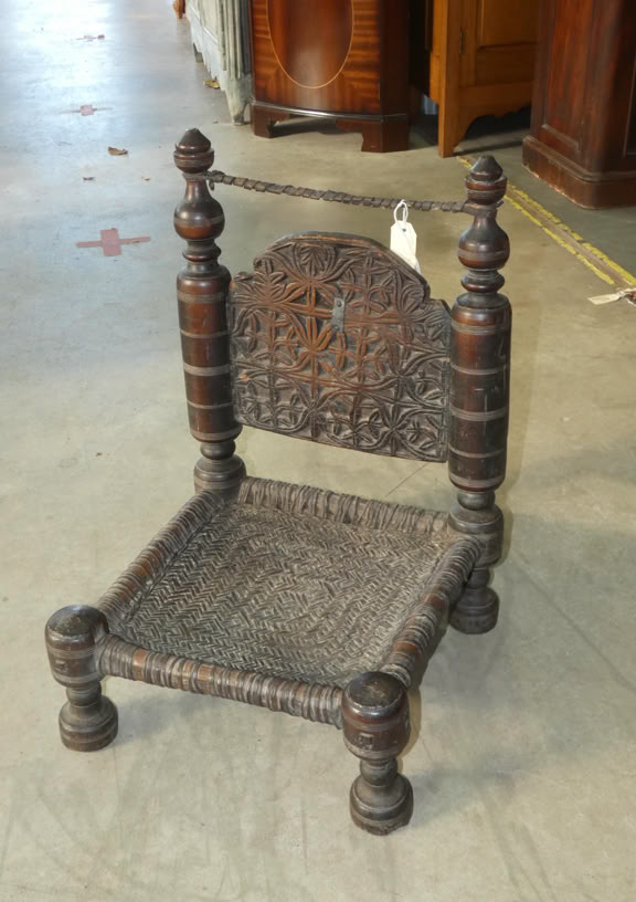 A small, intricately carved wooden chair with a woven seat and four short legs, featuring tall, decorative back posts.
